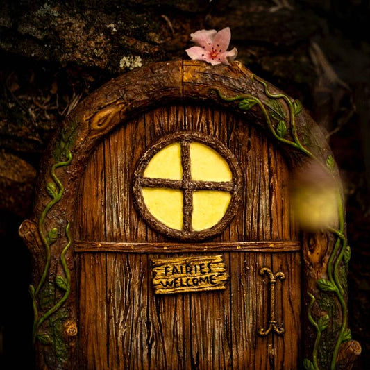 A whimsical wooden fairy door with a round window and a sign that reads 'Fairies Welcome'. It is adorned with a green vines around the door frame and a pink flower is visible on the left side.