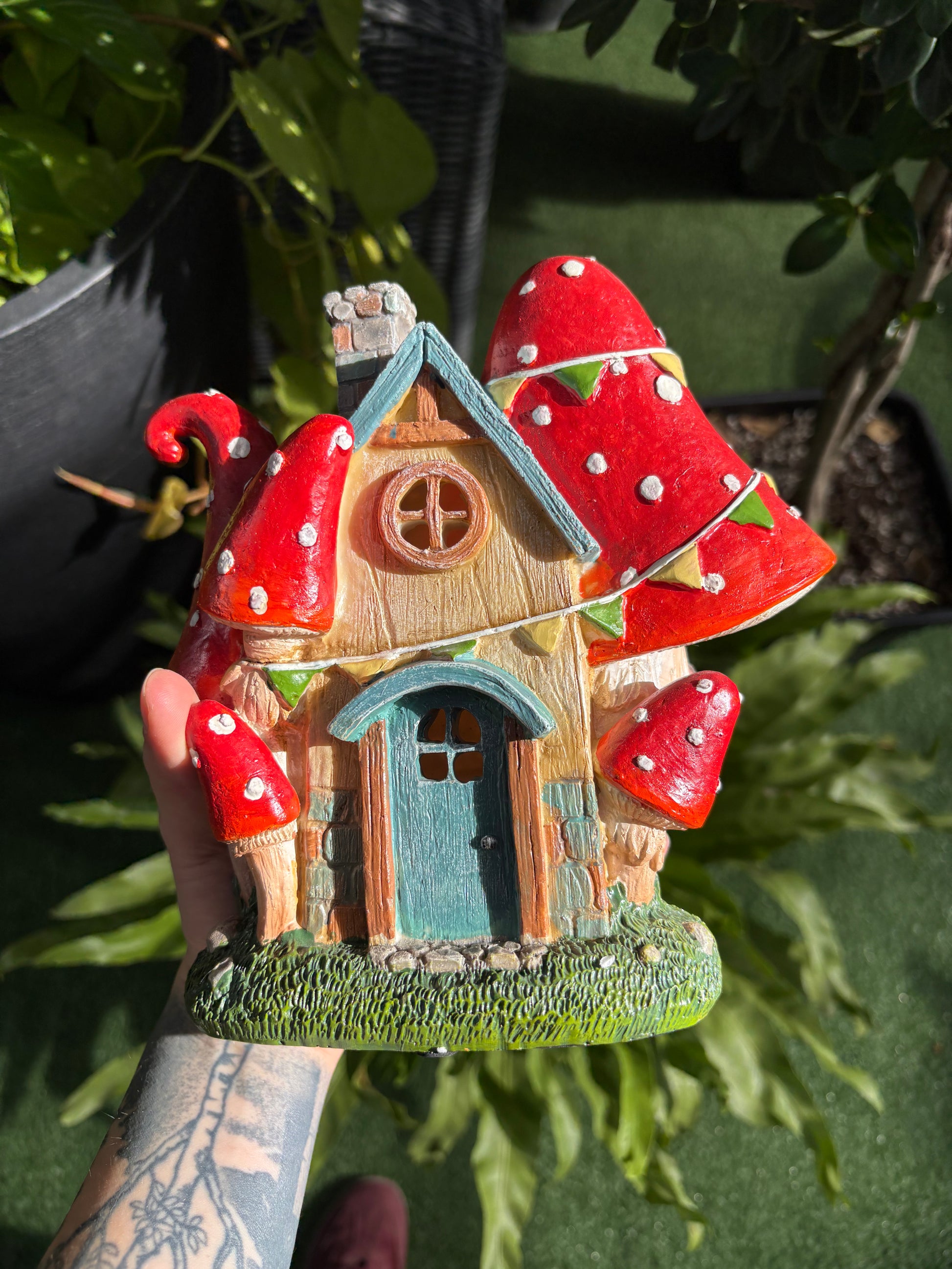 Hand holding a small, decorative fairy house with red roof and green base against a blurred natural background.