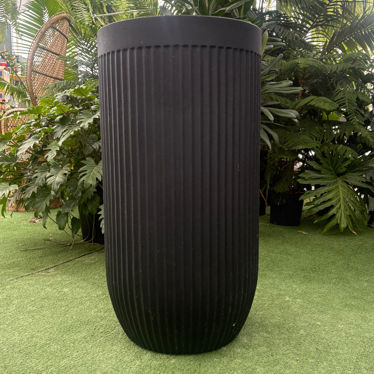 a stand alone matte black flute round tall black planter resting on a grass like surface in a greenhouse setting