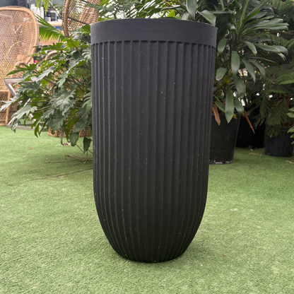 a stand alone matte black flute round tall black planter resting on a grass like surface in a greenhouse setting
