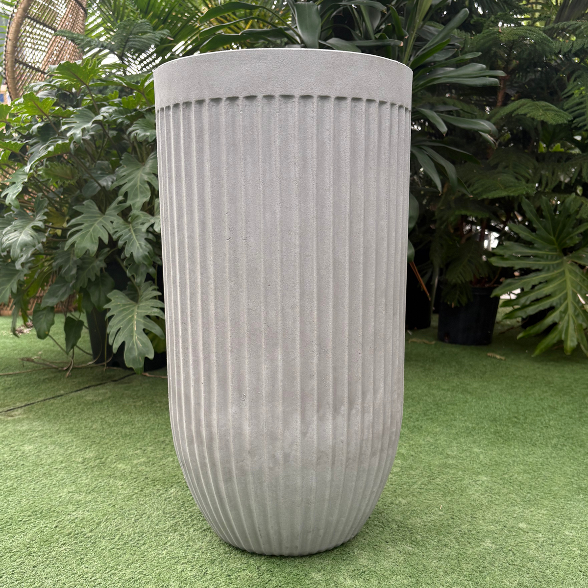a close up of a flute round burnt cement planter resting on a grass like setting in a greenhouse setting