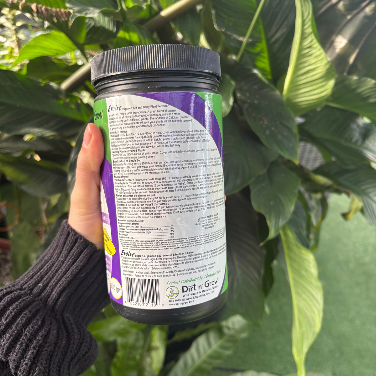 a person holding a black container showing the back of the product which has a labelled that describes the ingredients and product use with many tropical plants in the background