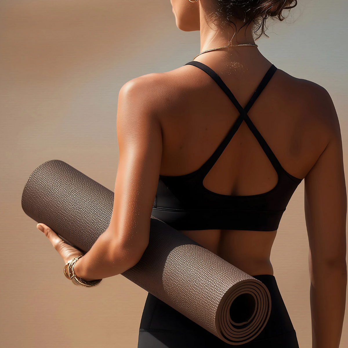 A woman shown from behind wearing a black sports bra and holding a rolled-up gray yoga mat against a warm beige background.