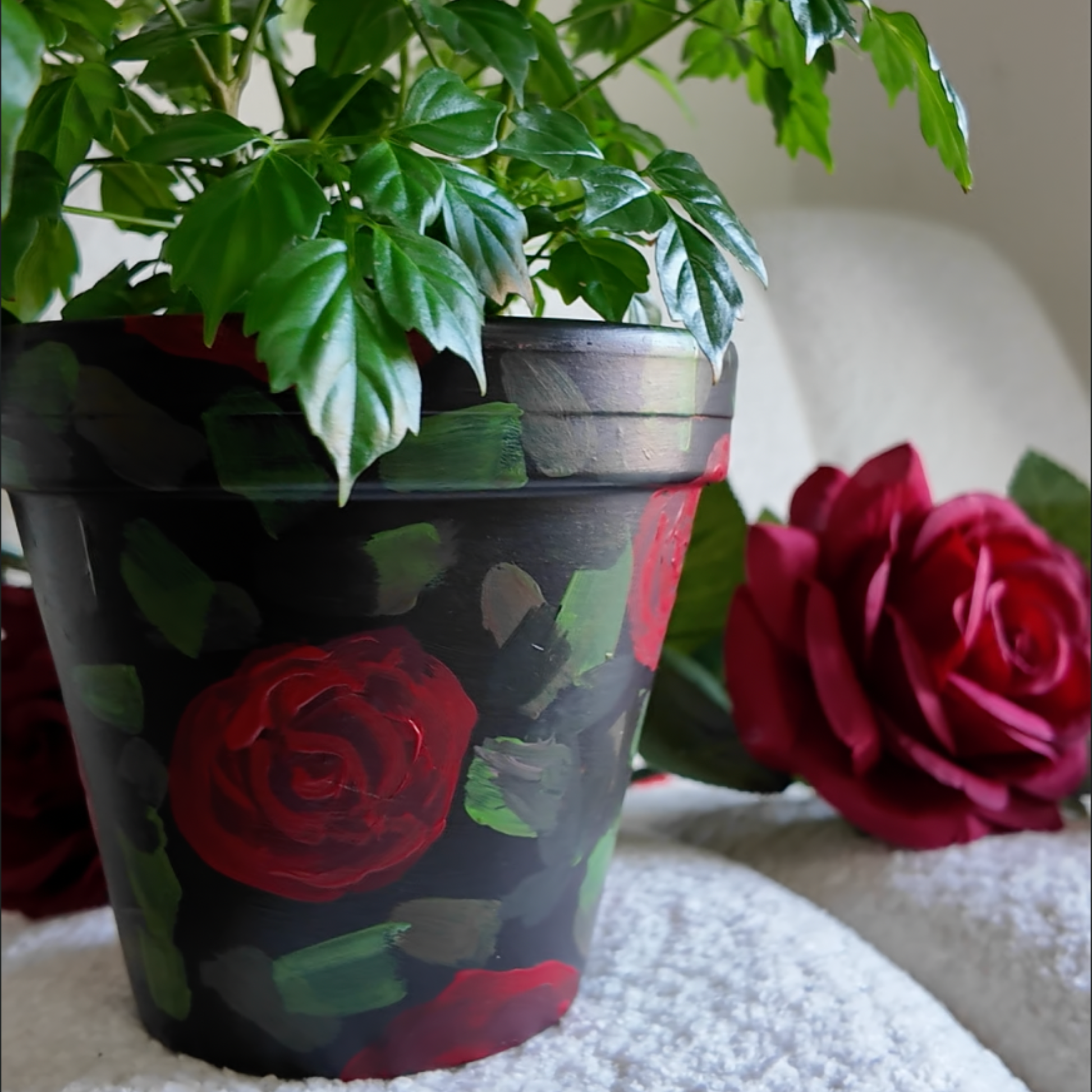 a picture of a plant pot that is hand painted with red roses and green leaves on a black background sitting on a white boucle chair with a red rose in the background