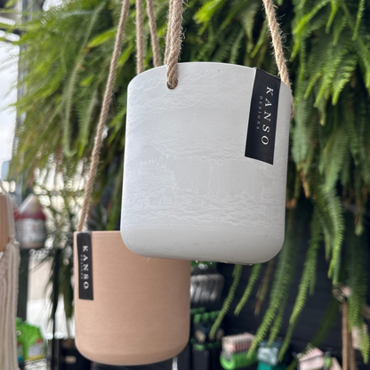 two four inch hanging pots one white and one a peach colour with a black label that reads Kanso designs hanging with twine like rope with many green ferns in the background