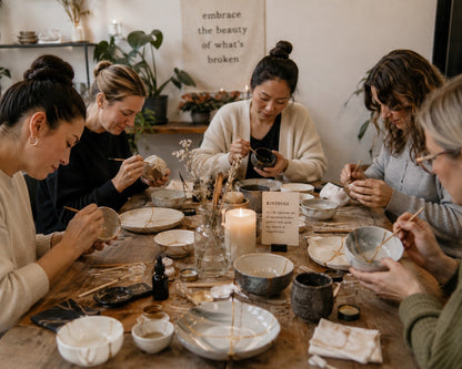 A group of five women gathered around a rustic wooden table, engaged in hand-painted pottery and ceramic crafting. The table is filled with various stoneware bowls, plates, brushes, and small bottles.