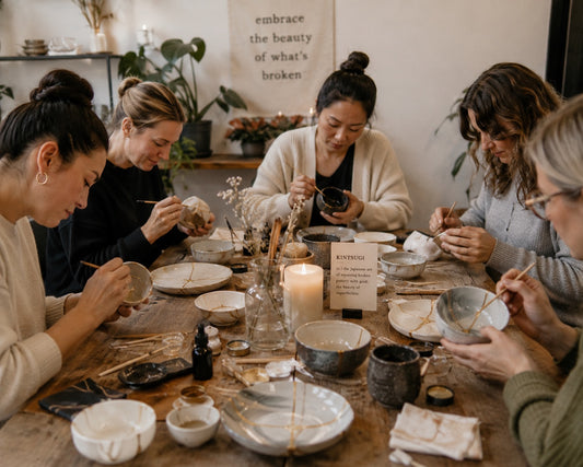 A group of five women gathered around a rustic wooden table, engaged in hand-painted pottery and ceramic crafting. The table is filled with various stoneware bowls, plates, brushes, and small bottles.