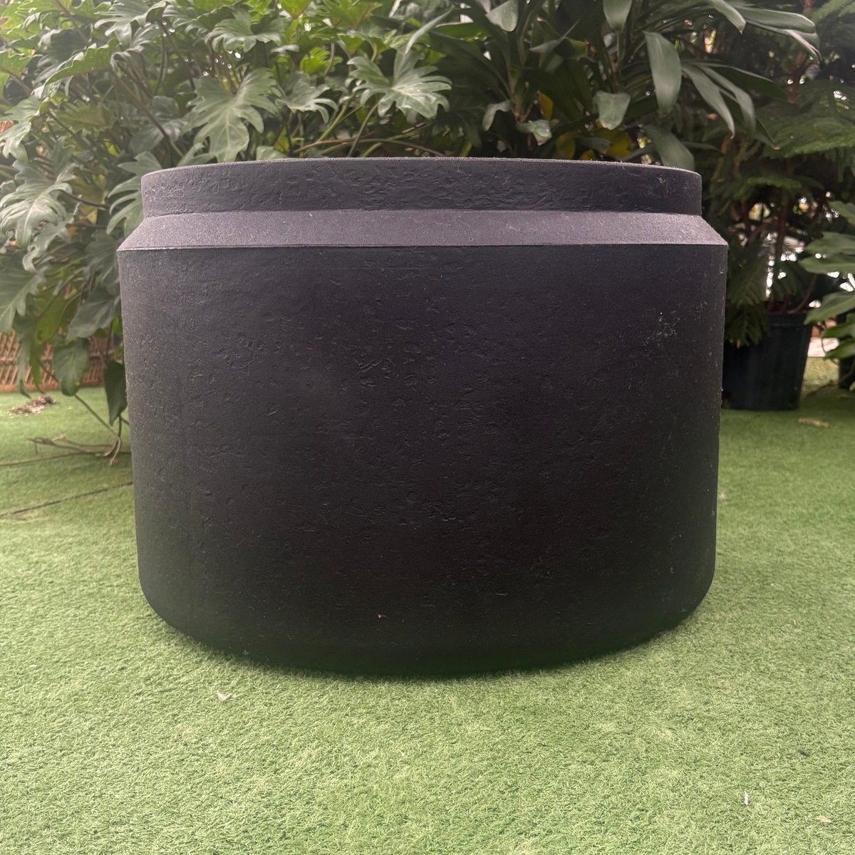 a close up of a matte black latte round planter resting on a grass like surface in a greenhouse setting