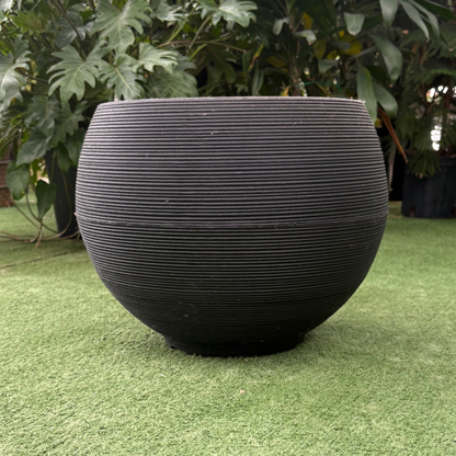a stand alone round matte black textured planter resting on a grass like surface in a greenhouse setting