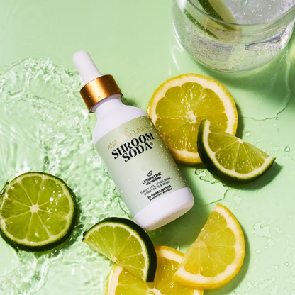 a white dropper bottle with a light green label that says shroom soda lemon lime, laying down next to many slices of both lemon and limes resting on a pale green surface
