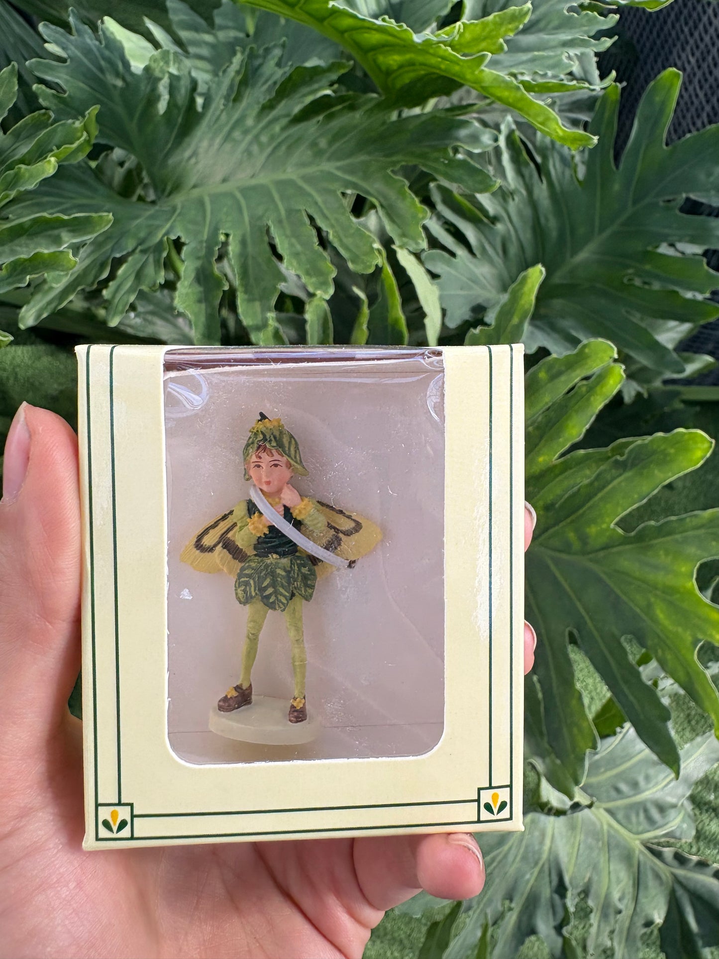Hand holding a small figurine in a decorative box with green leafy plants in the background