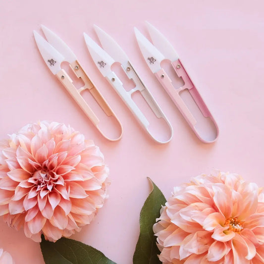 A set of mini plant snips with ceramic blades, in various pastel colors, displayed on a pink background with flowers.