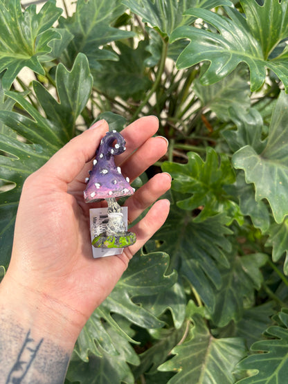 Hand holding a small purple and white decorative mini toadstool with a plant in the background