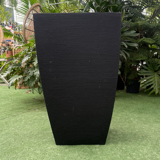 A tall, black rectangular planter with a textured finish, standing on artificial green grass in an outdoor garden setting with plants and wicker chairs in the background.