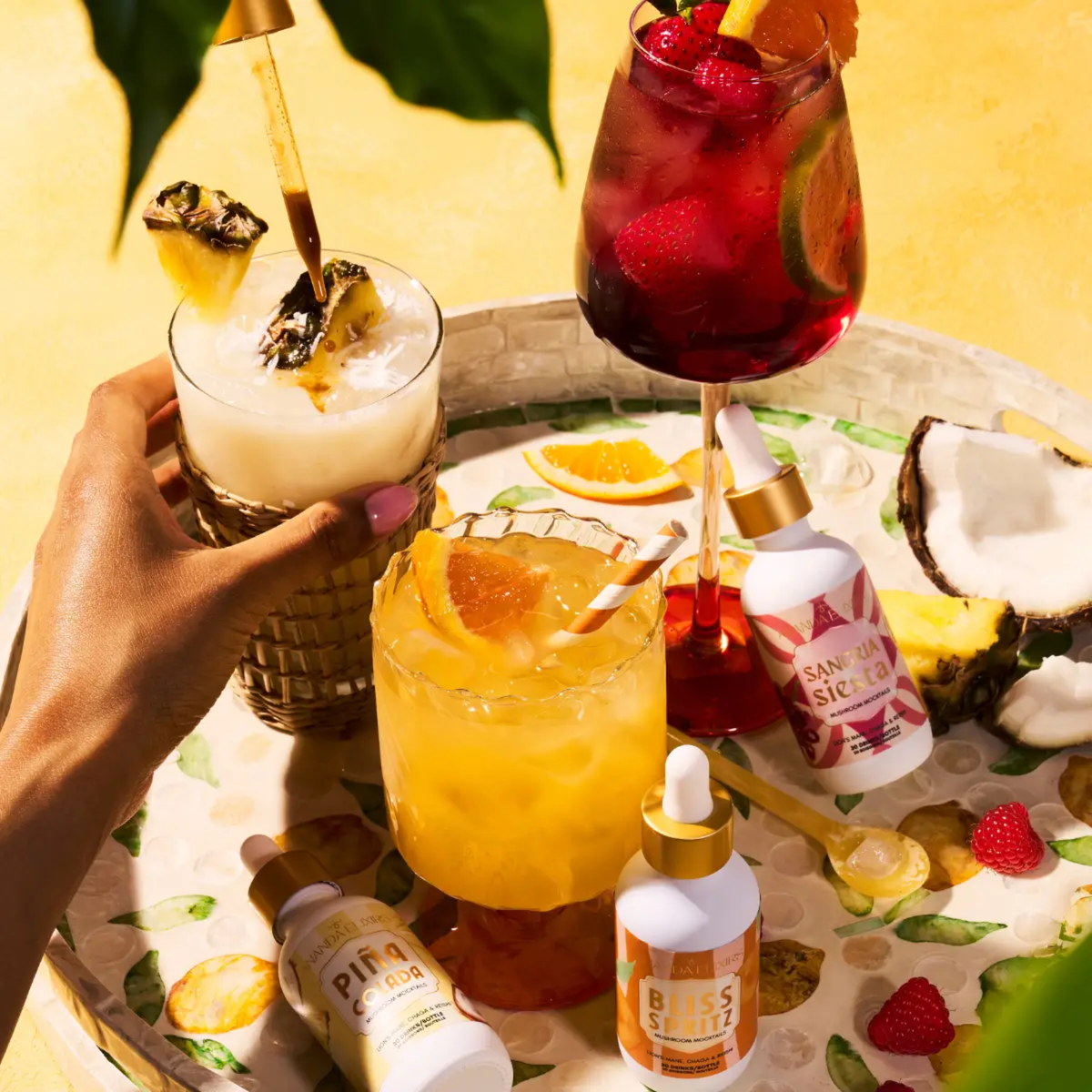 a tray of different cocktails with small droplet bottles next to their same cocktail types with fruit scattered on the tray with a yellow background and a person holding one of the drinks