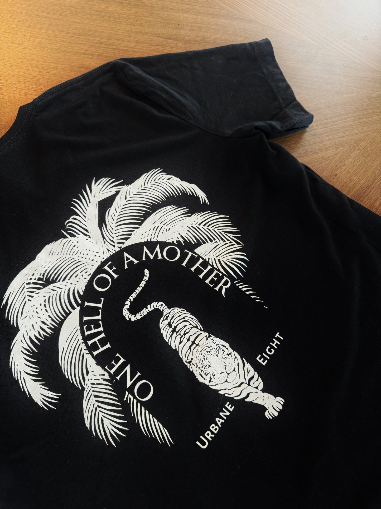 Black t-shirt with a white graphic of a tiger and palm trees, and text reading 'One Hell of a Mother' and 'Urban Eight'.