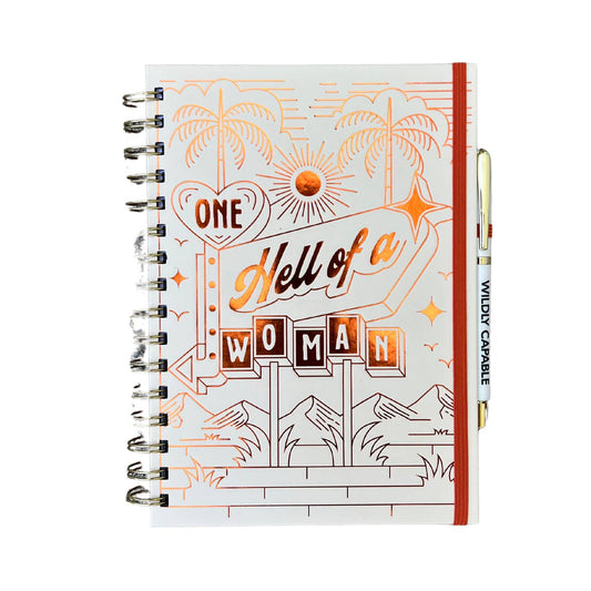A spiral-bound notebook with an orange and white cover featuring the text 'One Hell of a Woman' in stylized font, alongside a small illustration of palm trees.