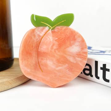 A hair clip shaped like a peach with a smooth pink color finish, placed on a wooden surface next to a bottle and a book.