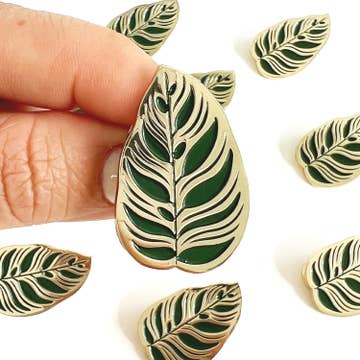 A collection of brooch pins shaped like green leaves on a white background, with one pin being held by a hand.
