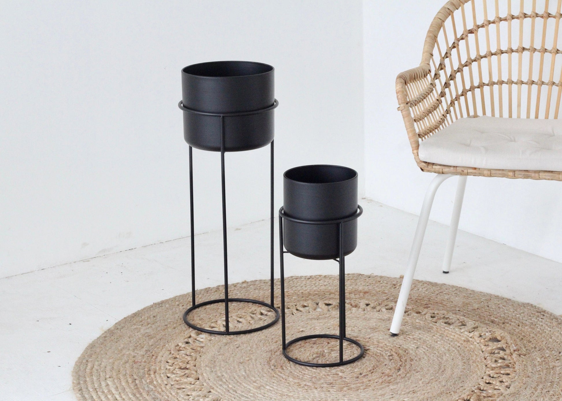 Two black metal plant stands on a jute rug, one tall and one short, with no plants in them, placed next to a wooden chair.