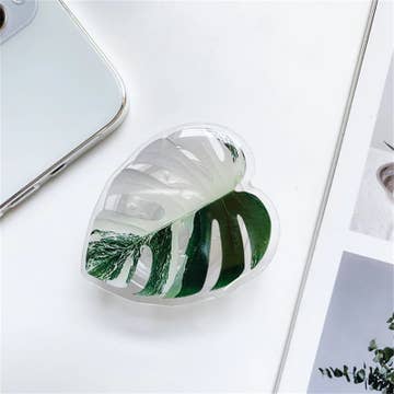 A heart-shaped, plant-themed phone holder placed on a flat surface next to a mobile phone and a small plant.