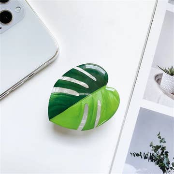 A green and white phone holder with a plant-themed design, placed on a surface next to a phone and a plant.