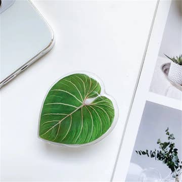 A green, heart-shaped phone holder with a leaf pattern, placed on a white surface next to a mobile phone and some decorative plants.