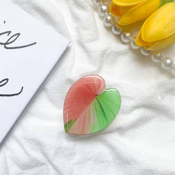 A heart-shaped phone holder with red and green colors placed on a white surface next to a pearl necklace and a note.