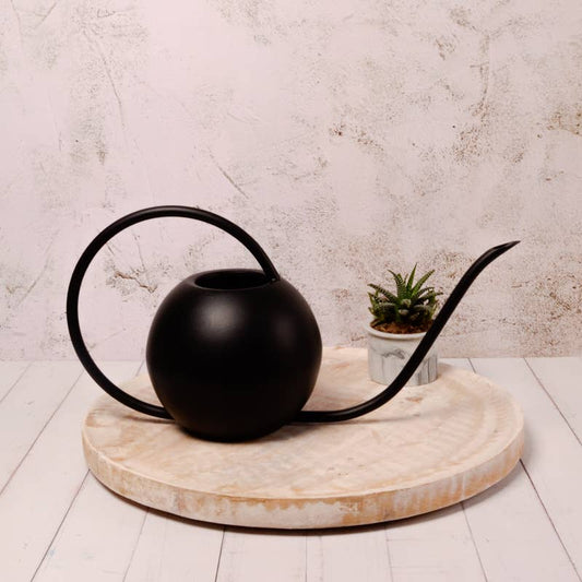 A black watering can with a long spout and a handle, placed on a round white wooden surface with a potted plant beside it.