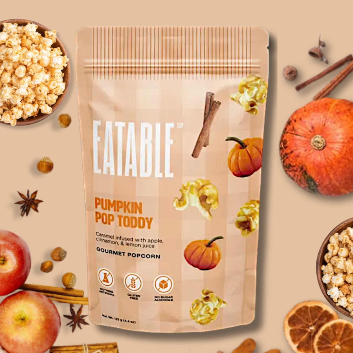 A stand-up pouch of Eatable Pumpkin Pop Toddgy gourmet popcorn, surrounded by autumn-themed ingredients like apples, cinnamon sticks, star anise, dried orange slices, and a small pumpkin on a warm beige background.