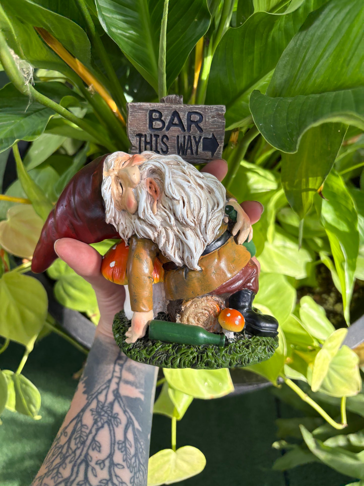Hand holding a figurine of a gnome with a beard and a sign saying 'Bar This Way' against a green leafy background.