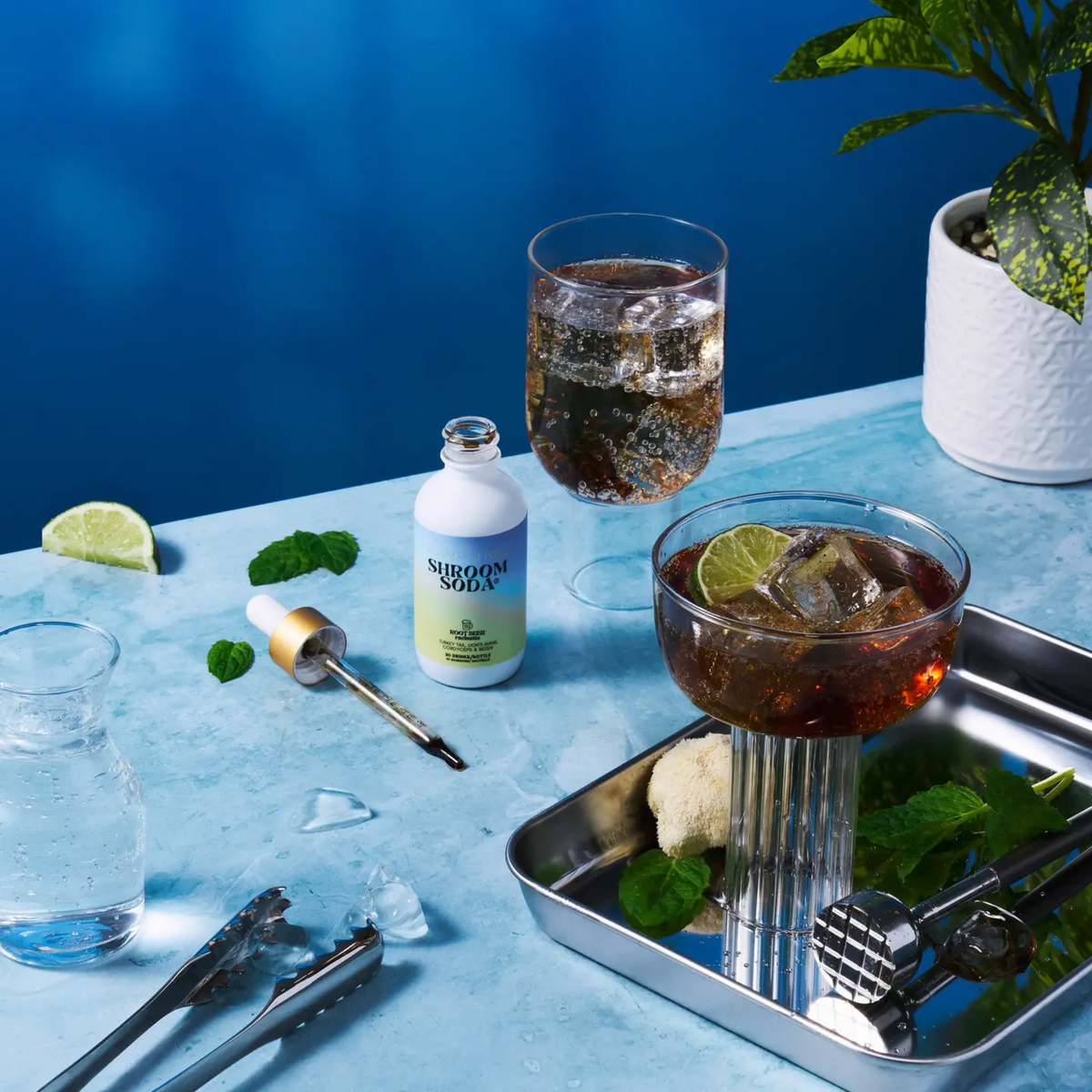 a white dropper bottle of shroom soda root beer with the lid off and the draping rising beside the bottle with two sodas in glasses on the same blue table with a blue background