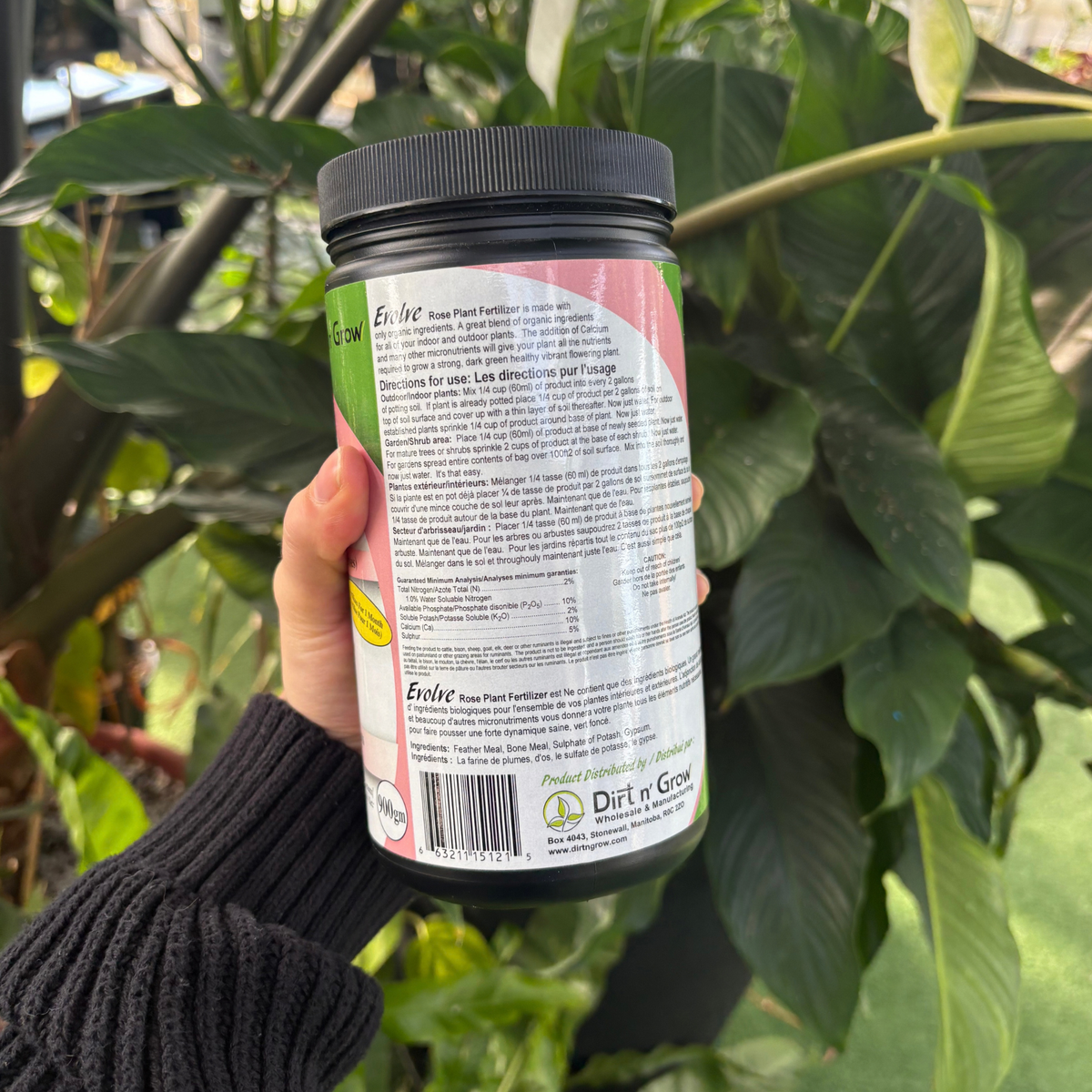 a person holding a black container with the back of the label facing forward displaying the product details, ingredients and use instructions with many tropical plants in the background