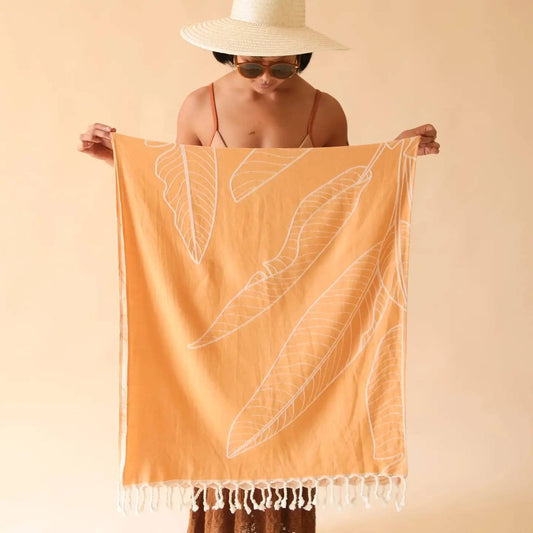 A person holding up an orange beach towel with white leaf patterns and fringes at the bottom.