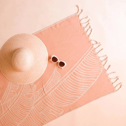 a pink sand resistant towel with a leaf pattern with sunglasses and a hat resting on it