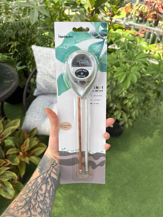 Hand holding a packaged 3 in 1 moisture meter against a green outdoor background