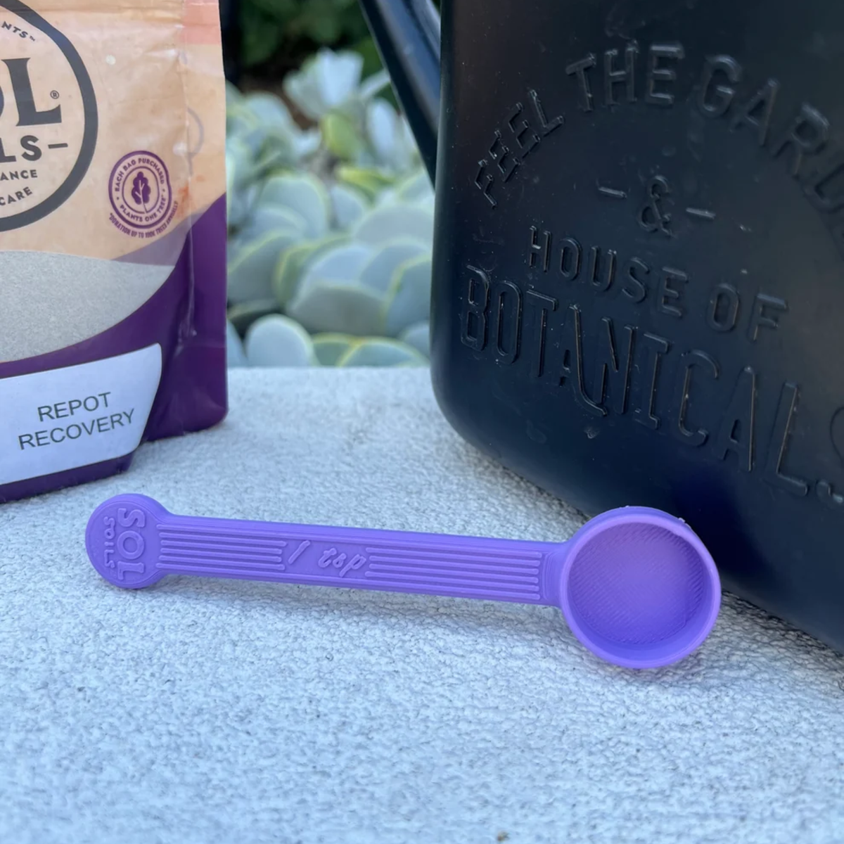 a purple teaspoon laying on a concrete surface with plants in the background