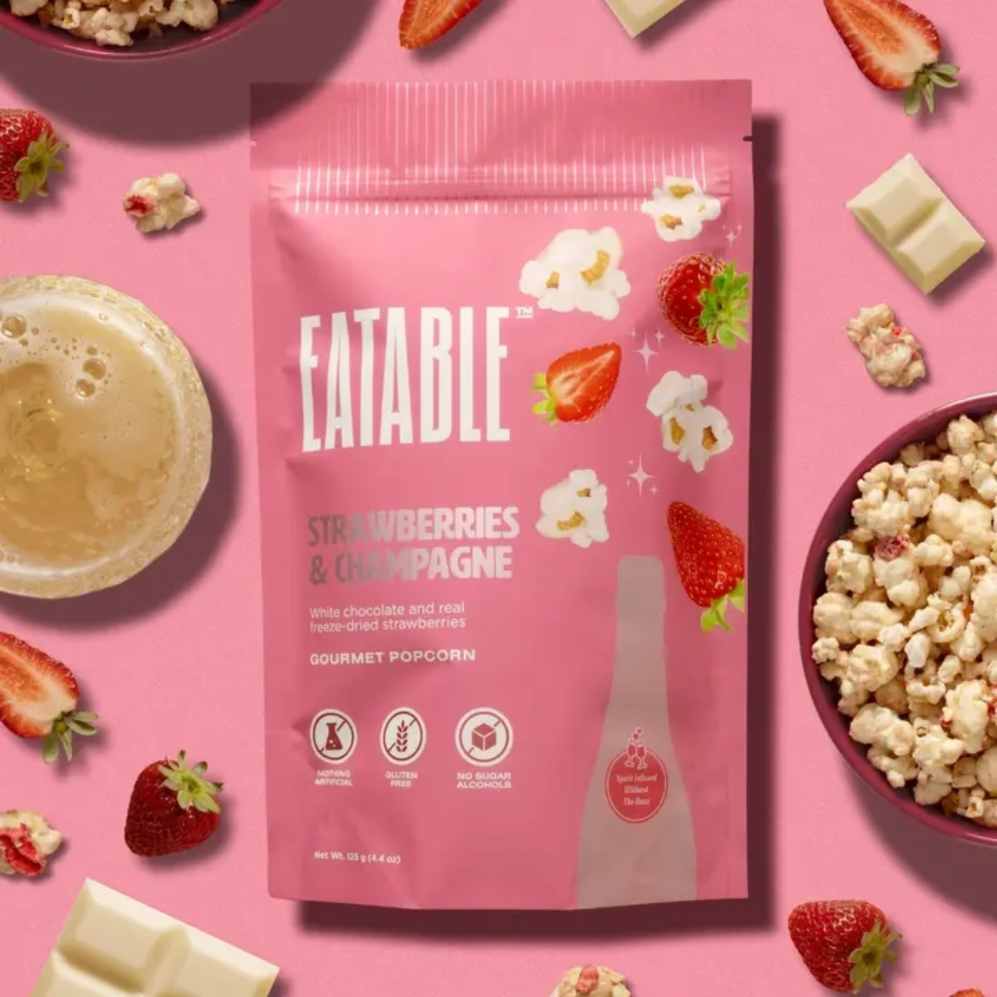 a pink bag of eatable strawberry and champagne white chocolate popcorn on a pink table with strawberries popcorn and champagne
