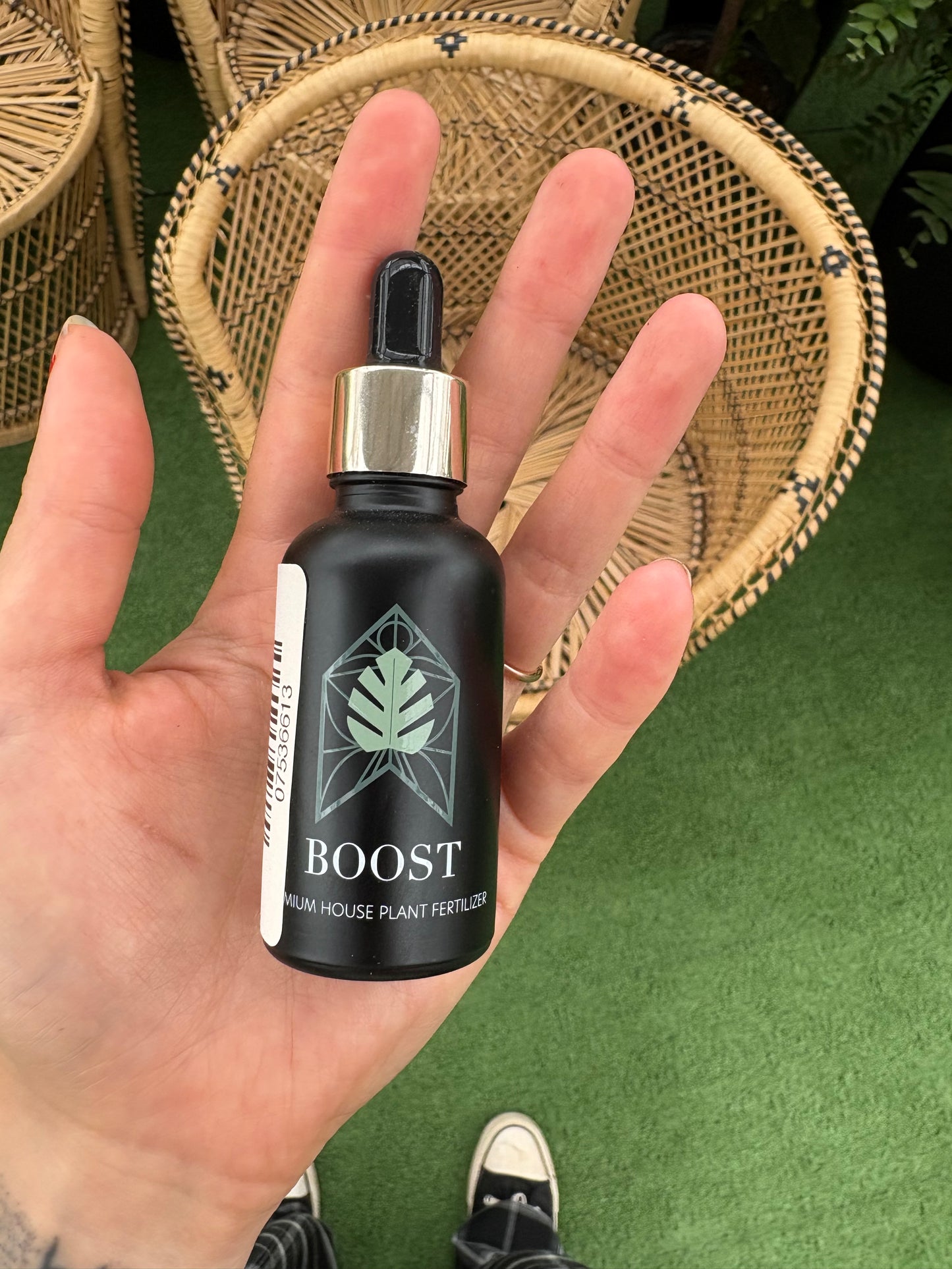 Hand holding a black plant care bottle labeled 'BOOST' on a green surface with a woven basket.