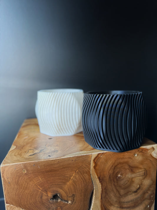 Two 3D printed pots, one white and one black, with a removable drip tray, placed on a wooden surface against a dark background.