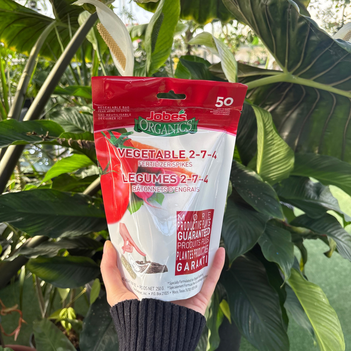 a person holding a white and red package that reads jones organics vegetable fertilizer spikes with many tropical plants in the background
