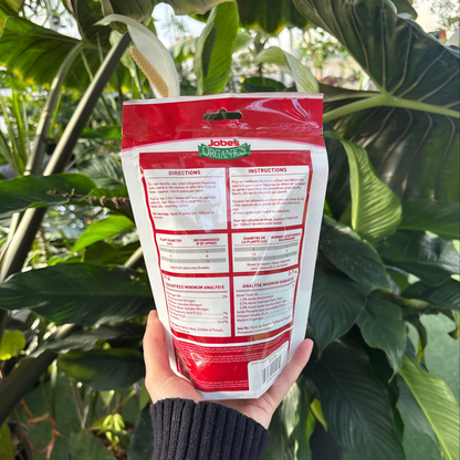 a person holding a white and red package of jobs organics vegetable fertilizer spikes displaying the back of the package which shows the ingredients and use instructions with many tropical plants in the background