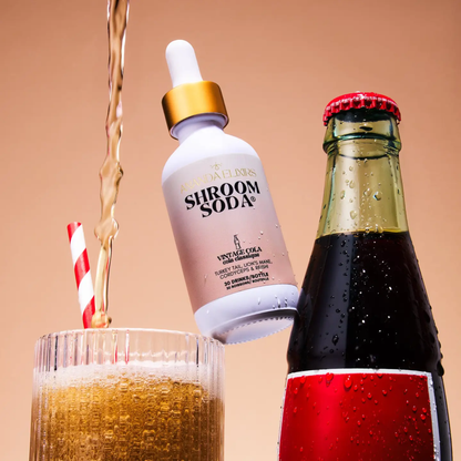 a white dropper bottle with a light pink label that reads shroom soda vintage cola with the bottle resting between a glass of cola and a vintage style cola bottle with a pinkish tan background