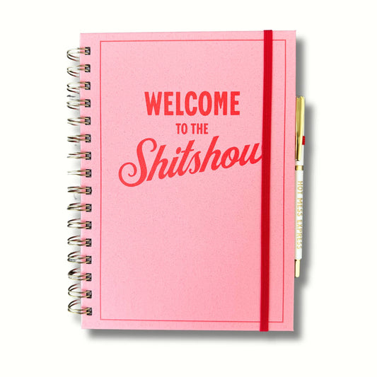 A pink notebook with the phrase 'Welcome to the Shit Show' printed in bold, red script.
