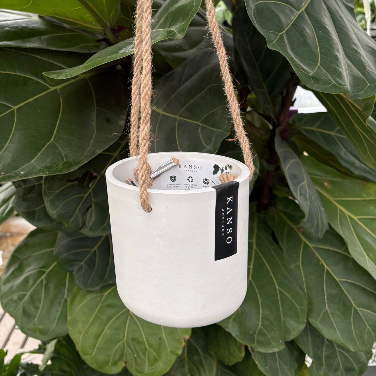 a white stone hanging four inch pot with a black label that read kano designs with the pot hung with twine like rope with a green tropical plant in the background